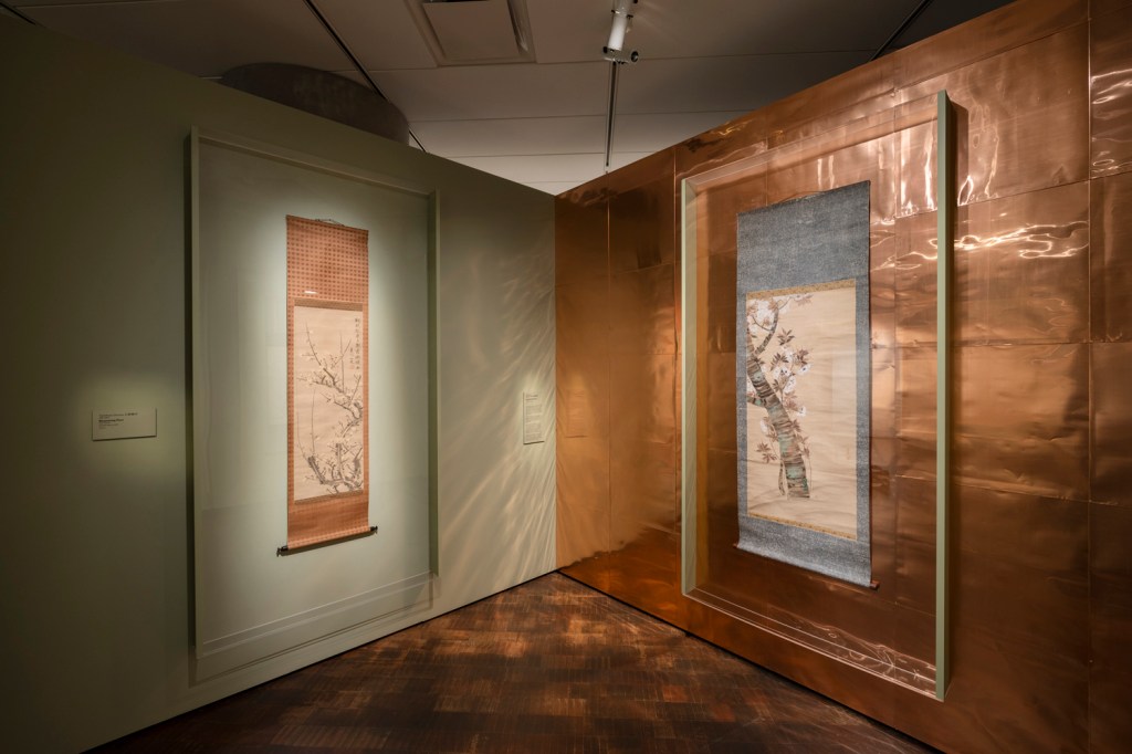 Two Japanese scrolls depicting botanical imagery in a small gallery space. One on a pale green wall, the other on a wall of bright copper.